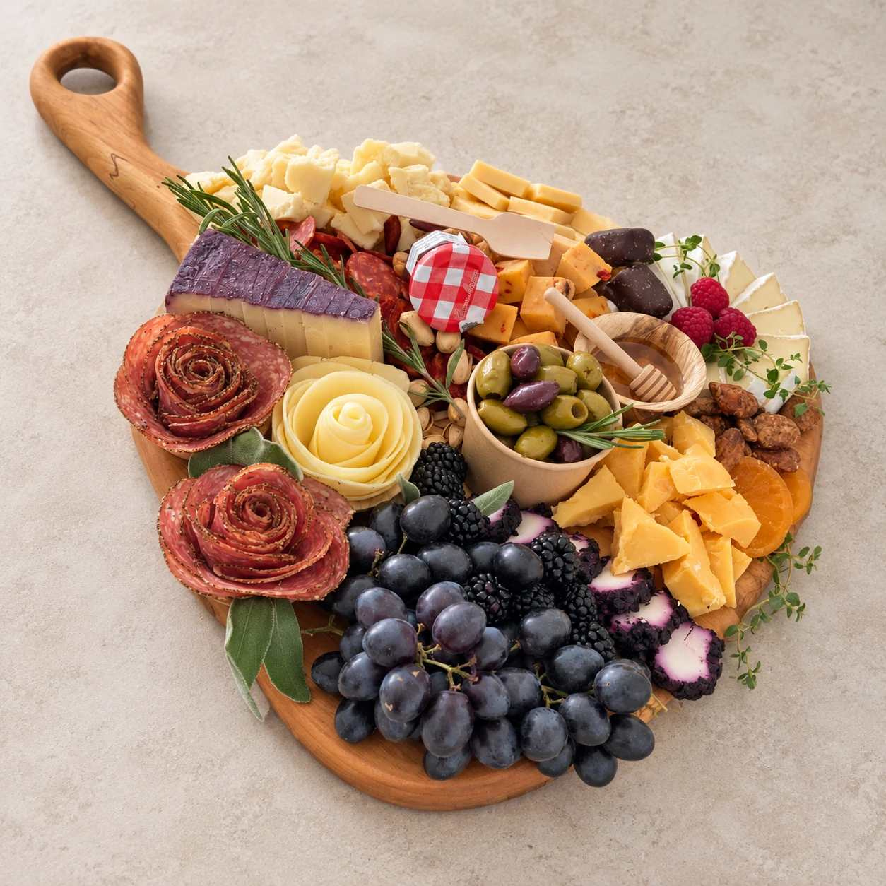 A handmade wooden serving board with a looped handle, loaded with cheeses, salami roses, grapes, olives and berries